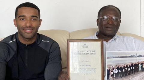 Nelson Shardey sits on his sofa holding his police commendation certificate, alongside his son Jacob