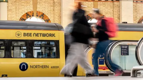 A northern train painted yellow with Bee Network written on, stationed at Piccadilly Station in Manchester. There is a blurred image of passengers walking in front. 