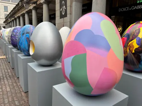 A row of large, colourful decorated eggs sit on grey plinths