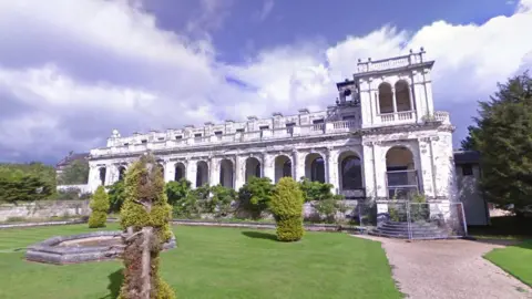 Google A derelict old hall, situated within an ornate garden with lawn in front. The building structure comprises a series of archways and balustrades.