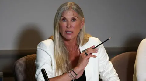 Sharron Davies, wearing a white jacket, sat behind a table at a press conference, with a microphone on the table