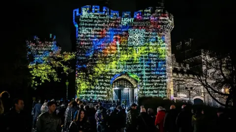 Robin Zahler The outside of Lancaster Castle at night lit up with mediaeval writing in white on blue, orange and yellow lights. There are lots of people milling about in front 