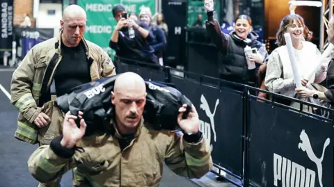 Kieran Thomas Kieran lunges whilst carrying sandbags. Andy stands behind - both wearing firefighting kit.