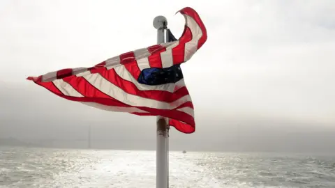 A US flag flutters in the wind. In front of it is open water. In the distance, through the fog can be seen the faint shadow of a long bridge.