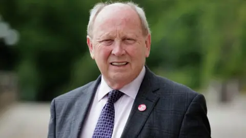 An older man looks into the camera smiling. he has grey hair at the side of his head and is bald on top. He is wearing a dark suit, white shirt and purple patterned tie. He is wearing a red badge that says 'no Irish sea border'.