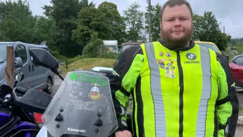 A man with a beard in a high vis jacket stands next to a motorbike in a car park with trees in the background. He has "BikerDan" written on the windshield of his motorbike. 
