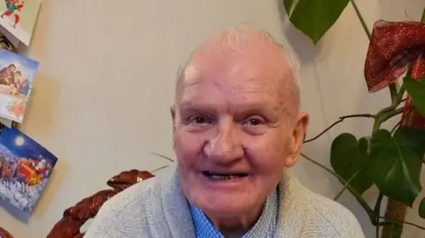 Craig family James Craig - an elderly man, sitting in a living room chair and wearing a jumper. Christmas cards are pinned to the wall behind him. 
