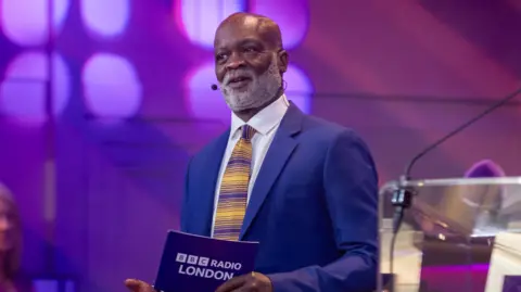 Eddie Nestor hosting a BBC Radio London event in a purple suit with a striped yellow and purple tie. He holding a programme card beside a lectern bearing the station’s logo.