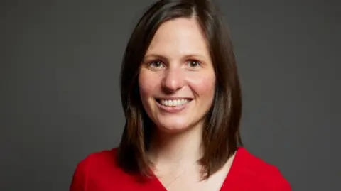 House of Commons/Laurie Noble/Parliament UK Official portrait of Sarah Smith MP who has shoulder length brown hair and wears a bright red jumper. She is smiling.