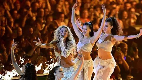 Getty Images Antigoni Buxton performs during a dress rehearsal for the second semi-final of the Eurovision Song Contest in Vienna. She has long, blonde curly hair and is wearing a white outfit that exposes her midriff. She's singing into a microphone that is in her left hand and her right arm is outstretched. Behind her are two dancers with dark hair in similar white outfits and in front of her are two dancers that are sat down. You can make out the silhouette of the crowd in the background.