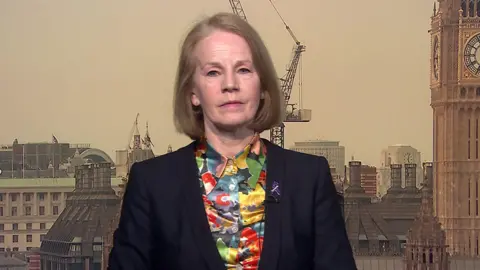 A picture of Baroness Bull, who has light brown hair and is wearing a dark jacket with a brightly coloured shirt underneath. A London skyline is in the background.
