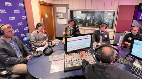 Simon Dedman/BBC Five young politicians sit in a radio studio in front of a presenter. They all wear headphones. The presenter has his back to the camera, wearing a black hoodie, and he reaches for one of the radio faders on a control panel.