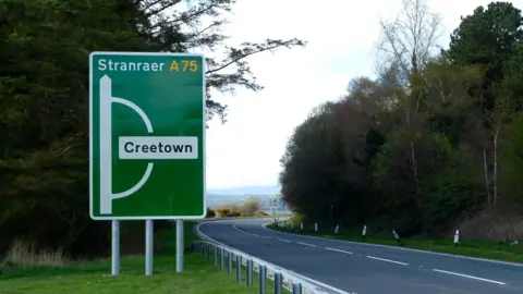 Anthony O'Neil A stretch of the A75 in south west Scotland, a road bending through the trees with a sign reading Creetown or straight on to Stranraer