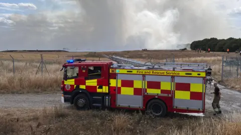 Simon Dedman/BBC A red and yellow fire engine is parked up on a dirt track, with dry brown grass either side. In the background is white smoke billowing.