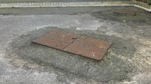 A double drain cover is higher than the surrounding road. There is a slope all the way around the drain cover to the road. It looks like a mini hill.