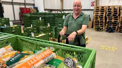 Volunteer, Paul Twyman, wears a green food bank polo shirt and stands in front of a pallet of pasta which has just been delivered. 