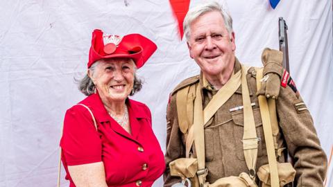An older woman wears a 1940s-style red dress and an older man wears a soldier's uniform