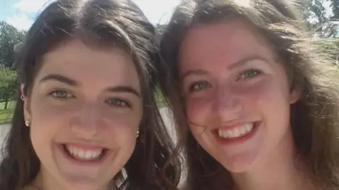Two sisters smile at the camera at a family wedding, both with long wavy hair
