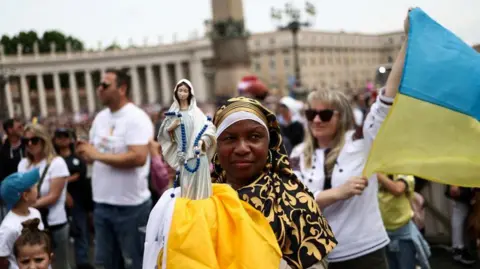 Reuters A woman in a vast crowd holds a small statue of the Virgin Mary, while behind her another woman holds a Ukraine flag