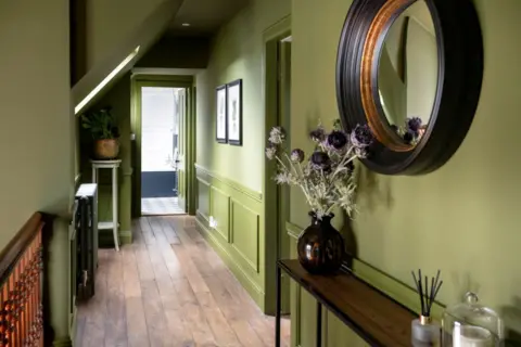 IWC Media Inside The Schoolhouse. The walls in the hall are olive green with a wooden floor. A large round mirror hangs on the wall and there is a vase on the sideboard.