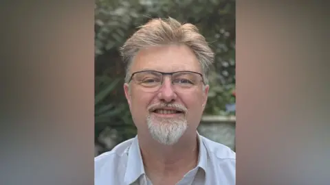 A man with glasses looking at the camera. He has light brown hair and a small beard which is grey. He is wearing a shirt and there is foliage behind him and possibly a large pot but it is a bit blurred.