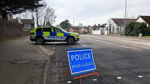 Scene in Dunmurry following a road traffic collision. There is a yellow, blue and white police vehicle to the left and a blue and white police road closed image