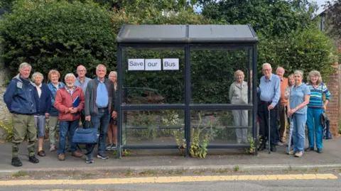 A gathering of people beside a bus shelter with a sign which reads Save our bus on it