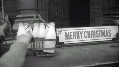 A black and white image of a hand on the left hand side sitting down a milkman's basket with around 7 bottles of milk in it.  On the right hand side there is a sign that says "MERRY CHRISTMAS"