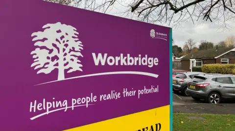 SAM READ/BBC A purple sign which features a tree motif and reads: Workbridge: Helping people realise their potential 