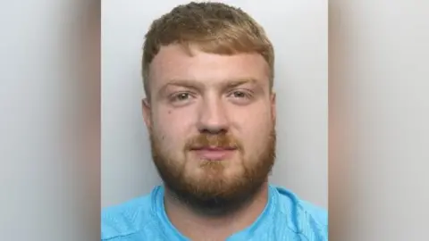 South Yorkshire Police A mugshot of Kaine Hicks, who has strawberry blond hair and a full beard of the same colour. He is smiling at the camera.
