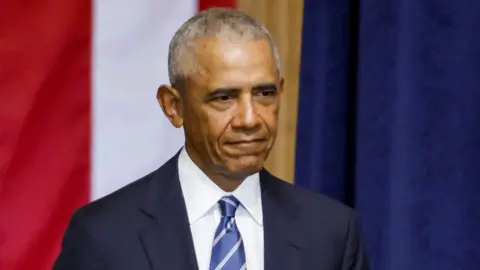 File photo shows a head and shoulders portrait of Obama looking to the right of camera while wearing a suit and tie against a red, white and blue background, at the Inauguration of Trump in January 2025.