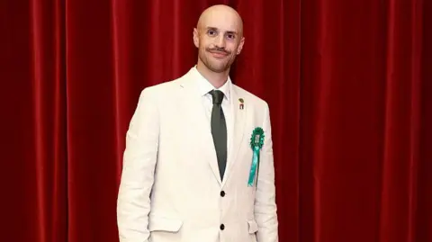 Getty Images A man in a cream linen suit, wearing a green rosette and green tie, stands in front of a red theatre curtain. 