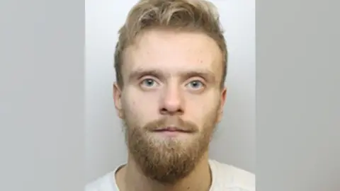 Mugshot of Ryan Scuffil, a 28-year-old man with pale skin, blue eyes, and blond hair. He has a blond moustache and short beard, and is wearing a white t-shirt.