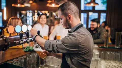 Getty Images A bartender, with short black hair and a black stubble, wearing a grey shirt, standing behind a bar. He is pulling a pint of beer. There are four people in the background, which is blurred.