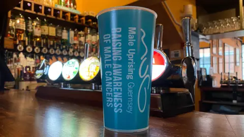 A blue reuseable plastic cup sits on a wooden bar top next to the beer and cider taps. There are bottles of spirits in the background.