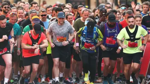A close-up picture of a people at the starting line as a previous year's run begins; one man is starting the timer on his watch.