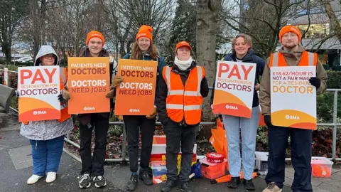 Five people standing outside Hull Royal Infirmary wearing orange beanie hats and holding placards. One reads: "Pay restoration for doctors". Another says: "Patients need doctors, doctors need jobs".