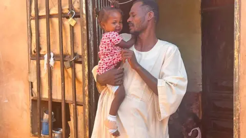 Ken Mungai/BBC Omnia, a little girl in a red flower print dress, is hugged by Uncle Abazaar Abdel Habib on a white plate. Her feet are bandages.