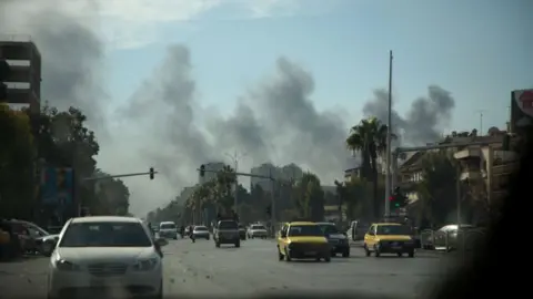 Smoke rising on the horizon of a street in Damascus, Syria. Cars are driving down the road. It is day time with a blue sky.