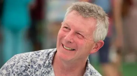 Independent Media News Stephen Moss wears a navy flowery shirt and has grey hair. He smiles and is turning to the left. The background is blurred in a green/blue tint.