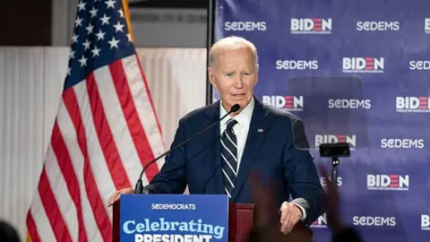 Joe Biden speaks at an event for South Carolina Democrats in February 2026. He stands at a podium that is fitted with a sign "Celebrating President Biden". An American flag hangs in the background.