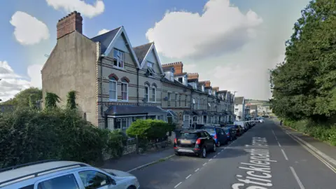 Sticklepath Terrace shows a long narrow road with a row of terraced houses on the left hand side. There are trees on the right hand side of the road. Several parked cars also line the lest of the road. It is a cloudy day. 