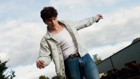 Fin Forster walking on train tracks wearing a cream jacket, white t-short and blue denim jeans with a brown belt. He has dark hair and has both arms stretched out to the sides to help with balance. There is a a long grey building behind him and trees