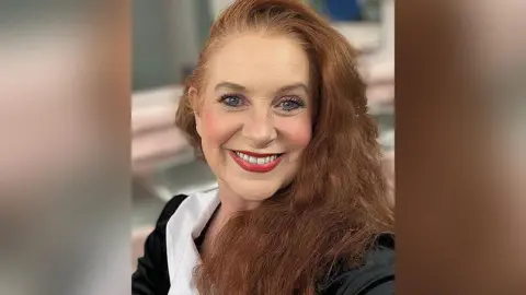 Cancer Research UK handout Close up of Jacqui Padden with long dark red auburn hair wearing a white collar and black satin style blazer. She is smiling. She is sitting in a sitting room which is blurred out in the background.