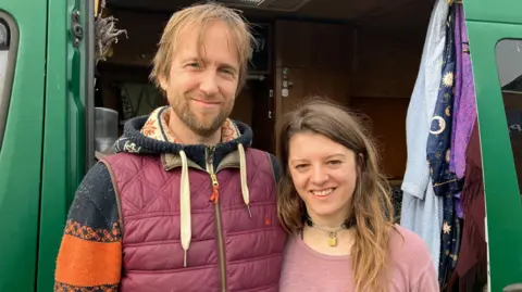 Luke Grossmith and his partner Jess Dane looking at the camera with their arms around each other in front of their green van. They are standing arm in arm and smiling. The door of the van is open with items of clothing hanging from the side of it.