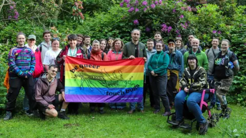 BBC / JOSH PARRY A large group gathered in a leafy park in Glasgow poses behind a rainbow flag reading “be queer touch bugs”, illustrated with insects. Participants stand and kneel closely together on grass, surrounded by dense greenery and blooming pink and purple flowers, with one person at the front crouching near the banner