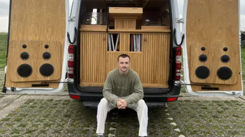 Lauren Carter/BBC A smiling Jack Lydon sits at the edge of his van, with the doors open and showing his DJ both. My Lydon is wearing white trousers and a green jumper, with his hands clasped together. Behind him you can see speakers attached to the inside of the van's doors and Japanese-inspired slatted cabinetry. 