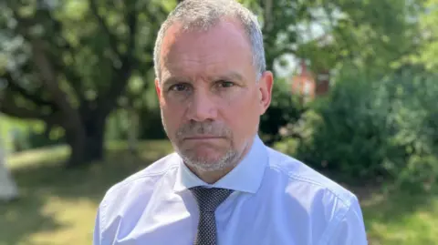 Adrian Leisk, who has short grey hair and a goatee beard, is dressed in a white shirt and tie. He is standing outdoors in a green park setting, with trees and blurred foliage in the background.
