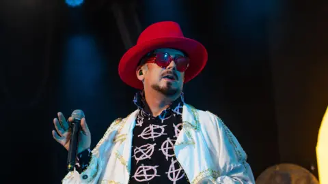 Boy George, wearing a red hat and grey shades, holds a microphone as he performs at the Henley Festival.