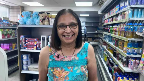 Kate Bradbrook/BBC A dark haired woman is smiling, behind her are lines of shop shelves. Some with soft drinks, others with dog treats, stuffed toys and miniature London red busses.